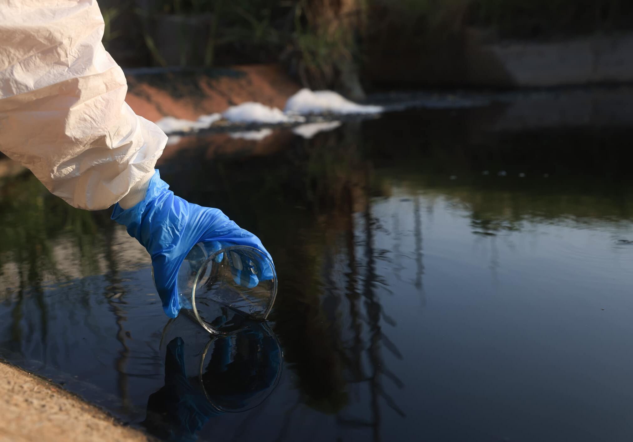 Close-up scientist hand with a white PPE suit and nitrile gloves collect sample waste water into a flask for laboratory water analysis, industrial and environmental concept